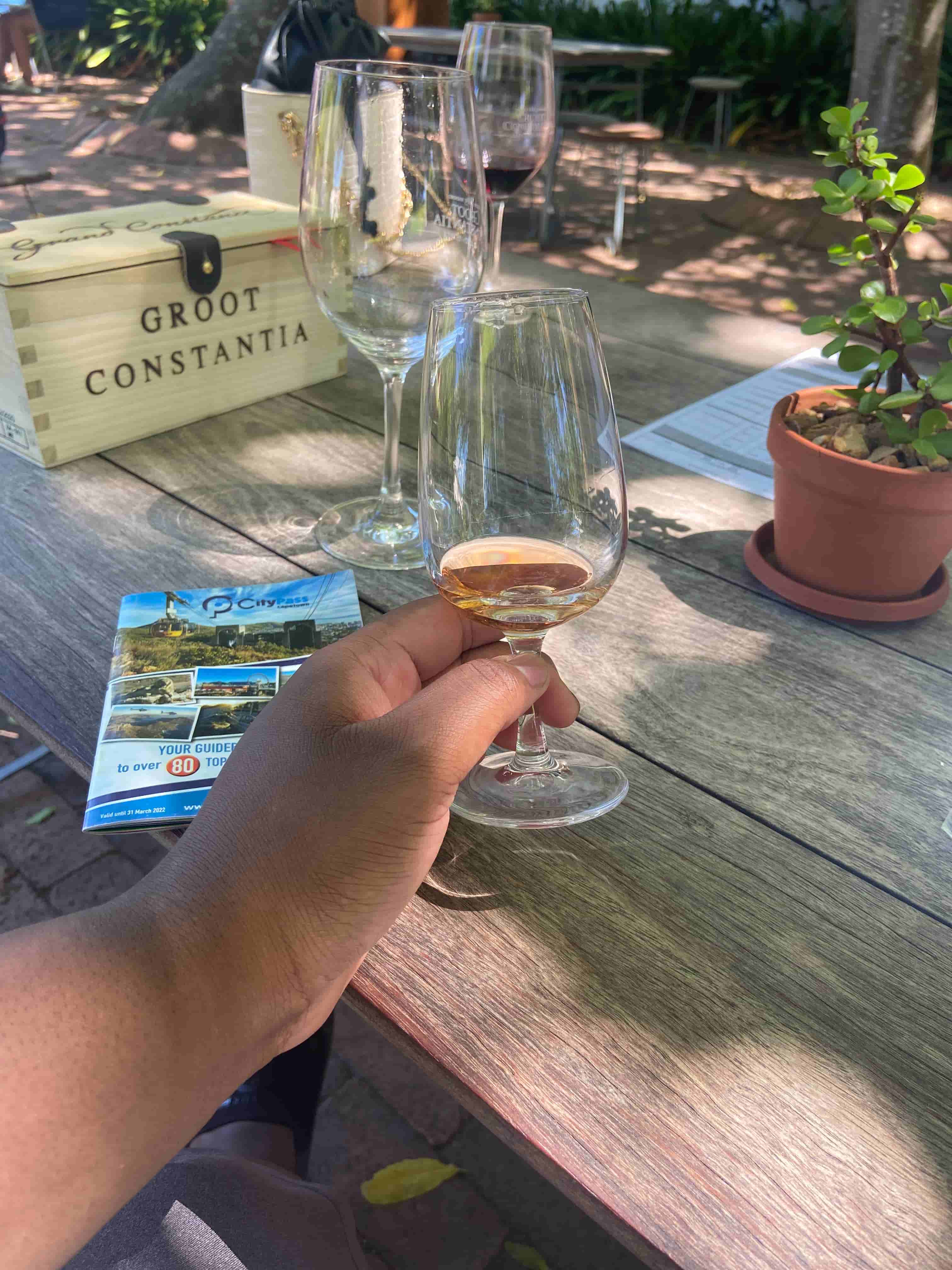 Holding out a glass of orange-pinkish wine on a park bench with a box saying Groot Constantia in the background