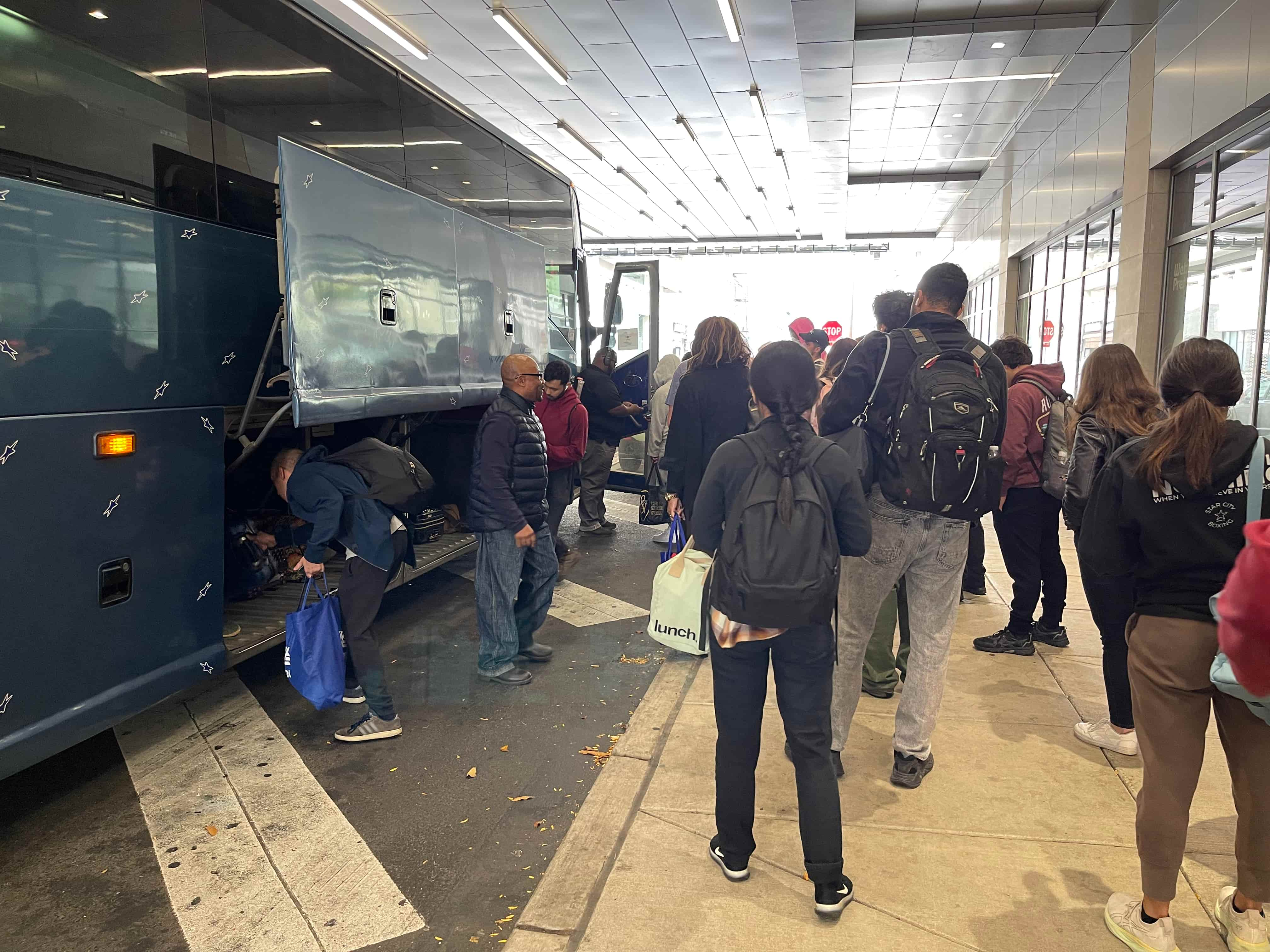 A bus parked by the side of a road in between a mall building with people boarding and placing their luggage