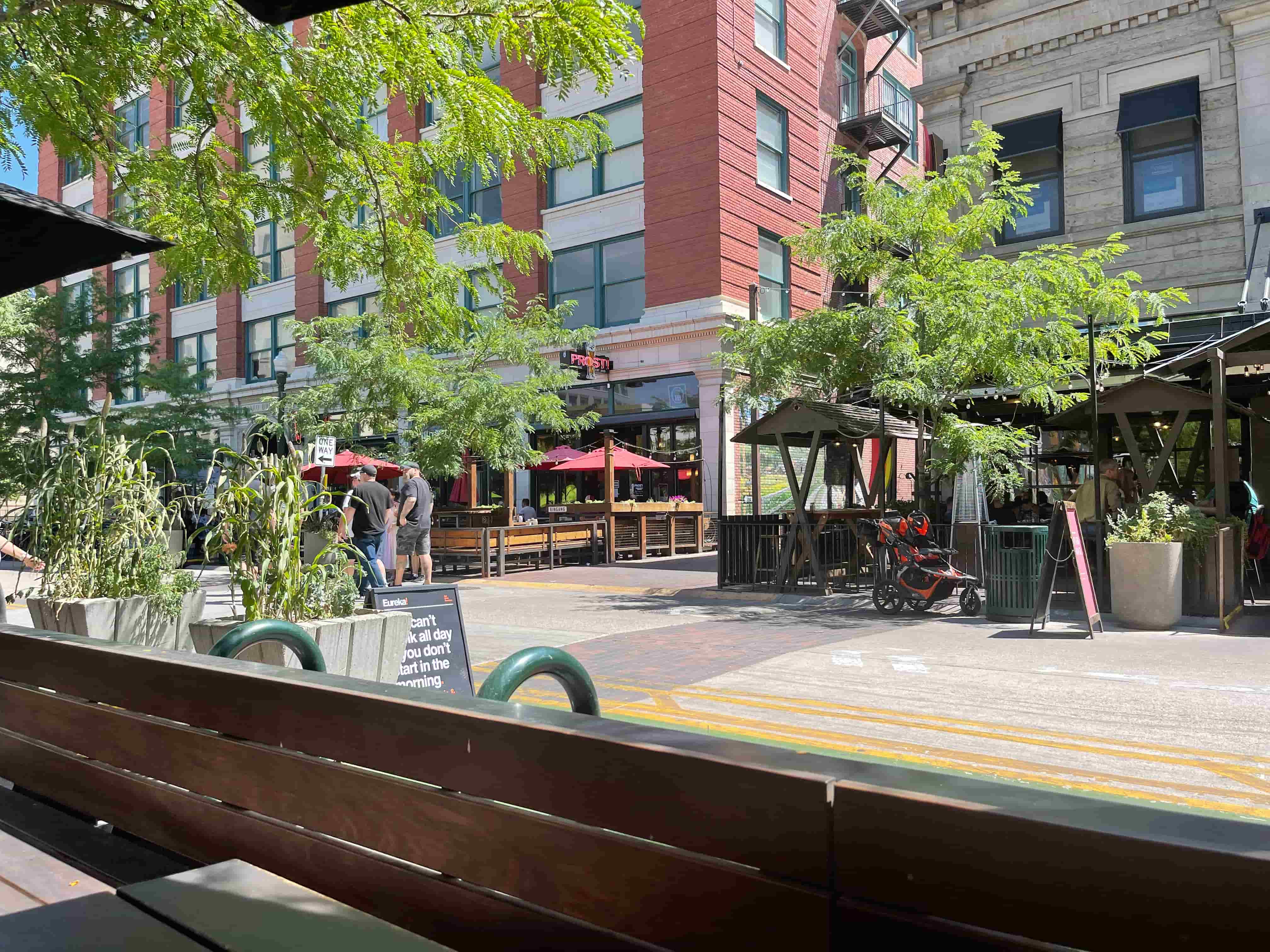 a photo of a pedestrianized street with some colorful medium sized apartment buildings, tasty foliage, outdoor dining seating and a couple people walking