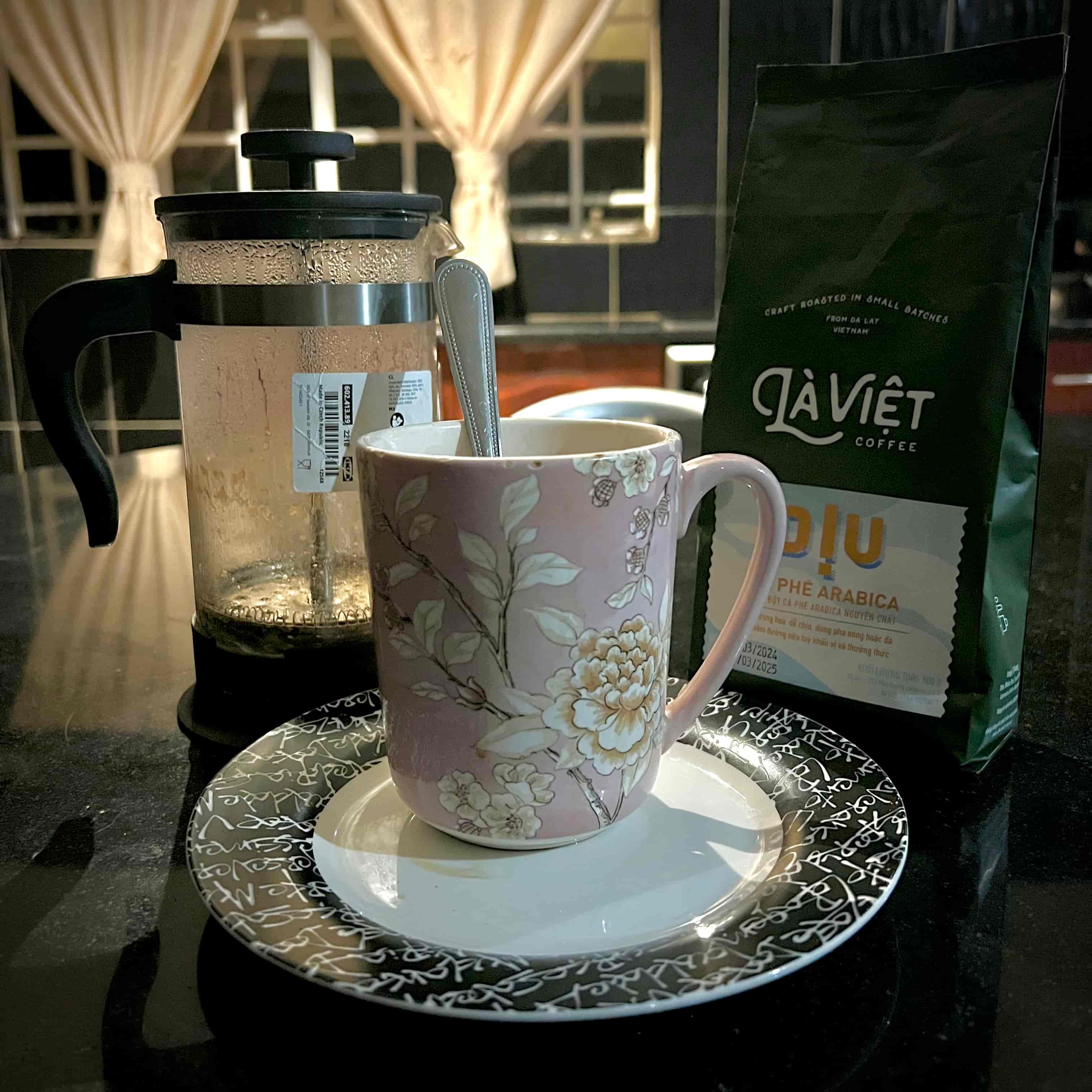 A flowery pink mug between a french press and a bag of Là Viêt coffee grounds